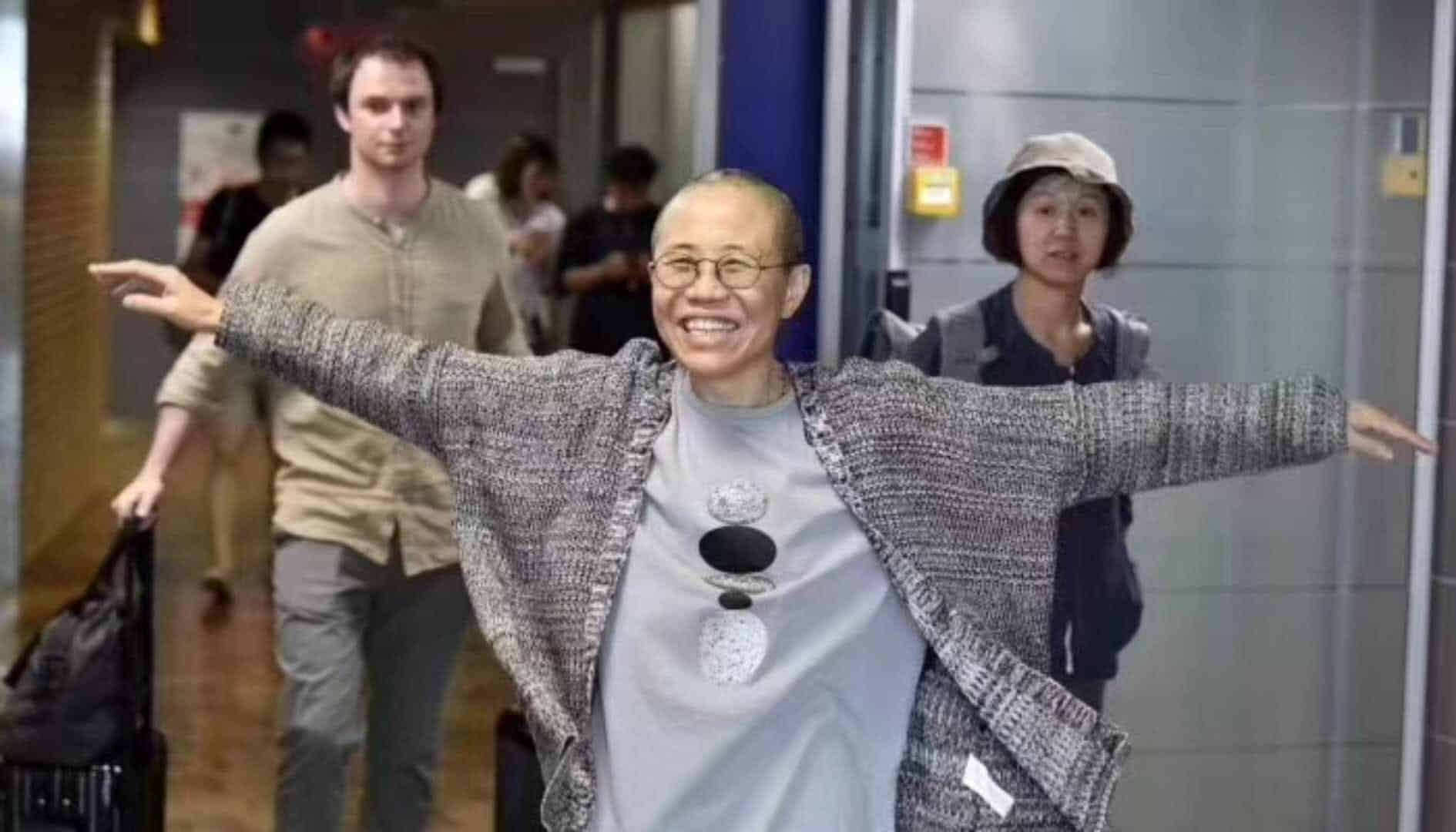 A person with short hair and glasses smiles broadly with their arms outstretched, walking through an airport corridor. Two people with luggage walk behind them.