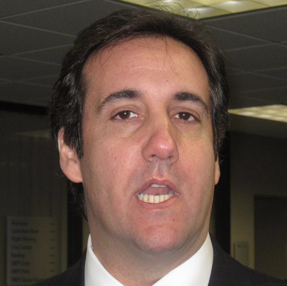 A man with dark hair wearing a suit and white shirt is speaking indoors under fluorescent lighting, with office ceiling tiles and a window in the background.