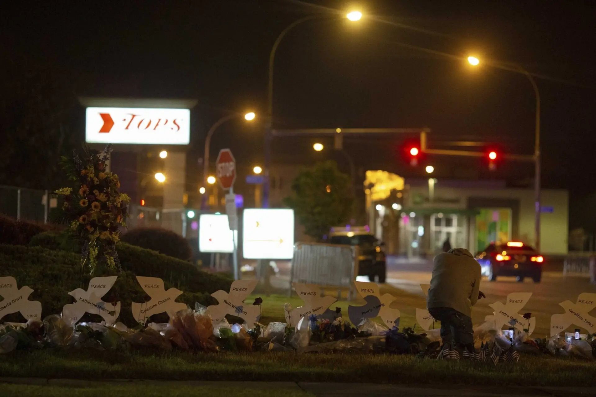 A person kneels at a nighttime memorial with white cross markers, flowers, and candles in front of a Tops store, illuminated by streetlights and neon signs in the background.