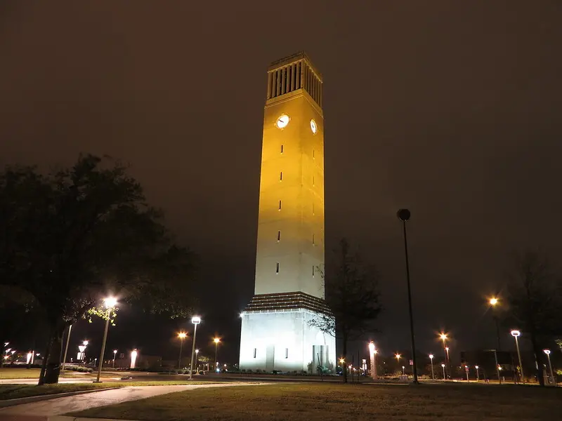 A tall clock tower illuminated at night stands in a park with trees and streetlights, under a cloudy dark sky.