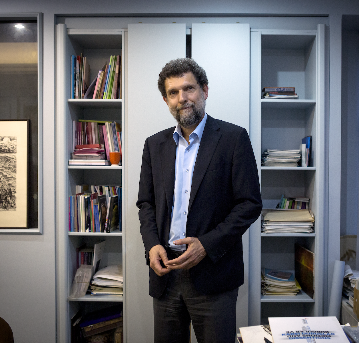 A man with curly hair and a beard stands in an office, wearing a dark suit and light blue shirt. He is in front of bookshelves filled with books, folders, and papers.