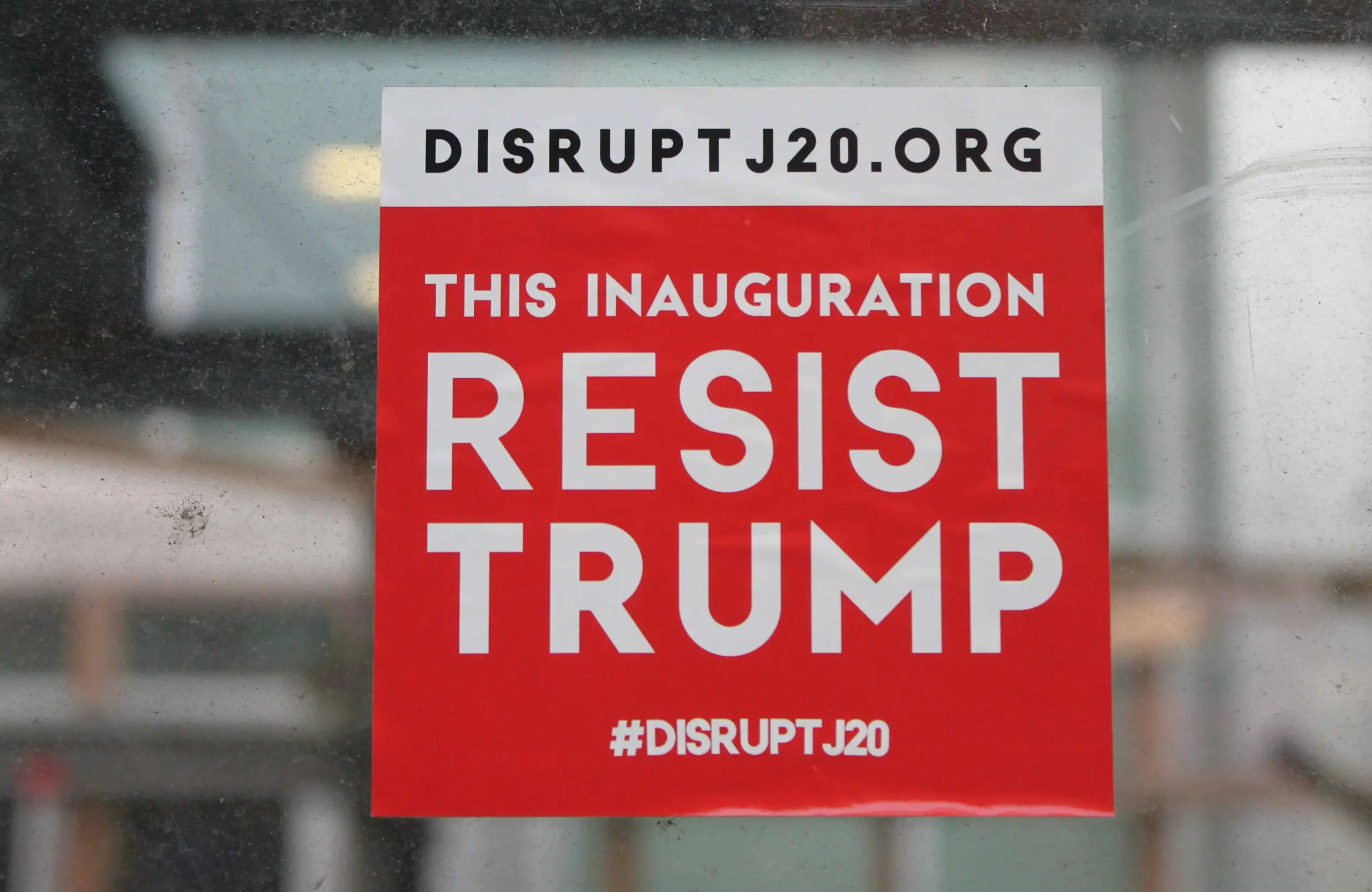 A red and white sign reads “DISRUPTJ20.ORG. THIS INAUGURATION RESIST TRUMP. #DISRUPTJ20” and is posted on a glass surface.