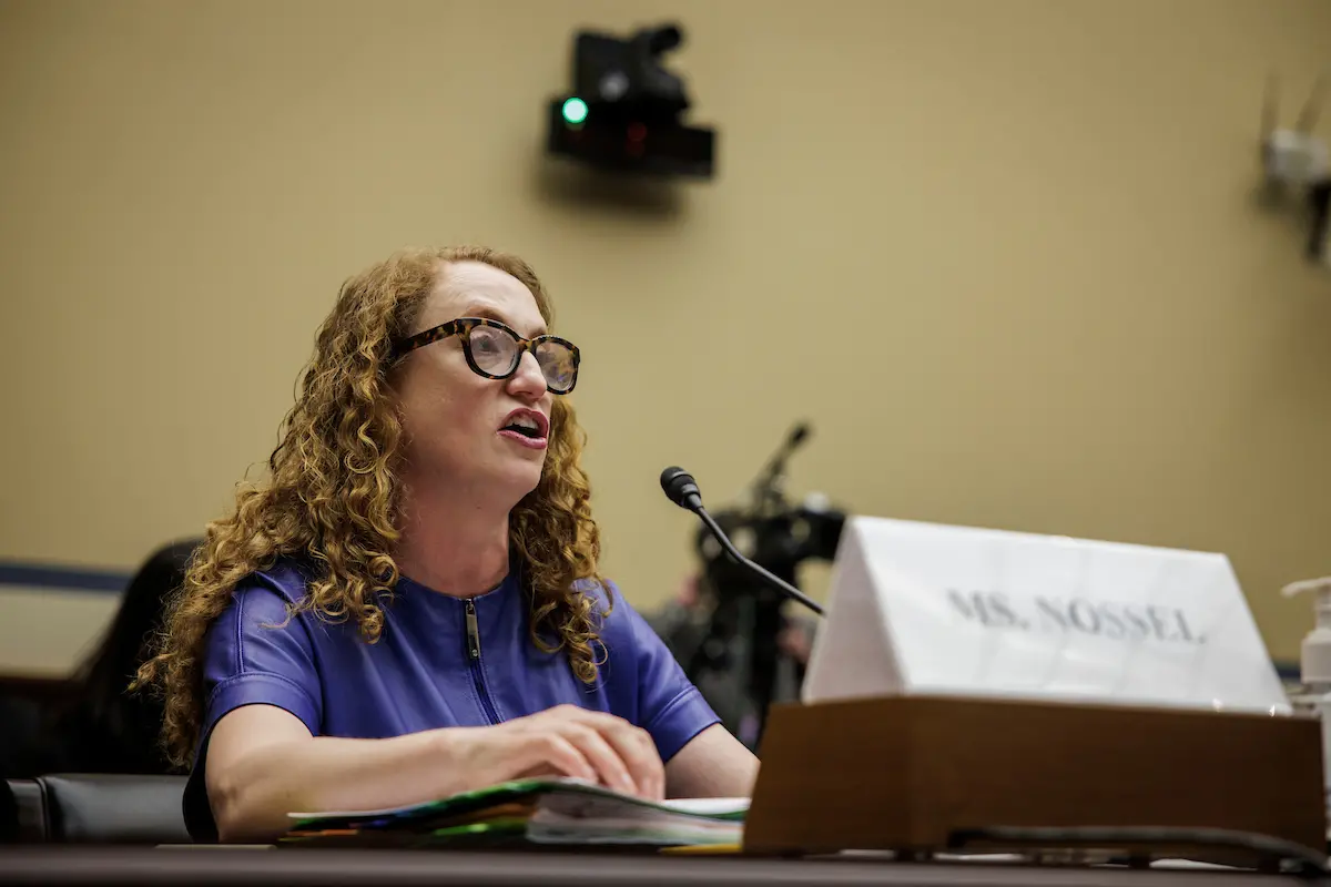 A woman with curly hair and glasses, wearing a blue top, speaks into a microphone at a hearing. A nameplate reading Ms. Nossel is in front of her on the desk, along with documents and a sanitizer bottle.