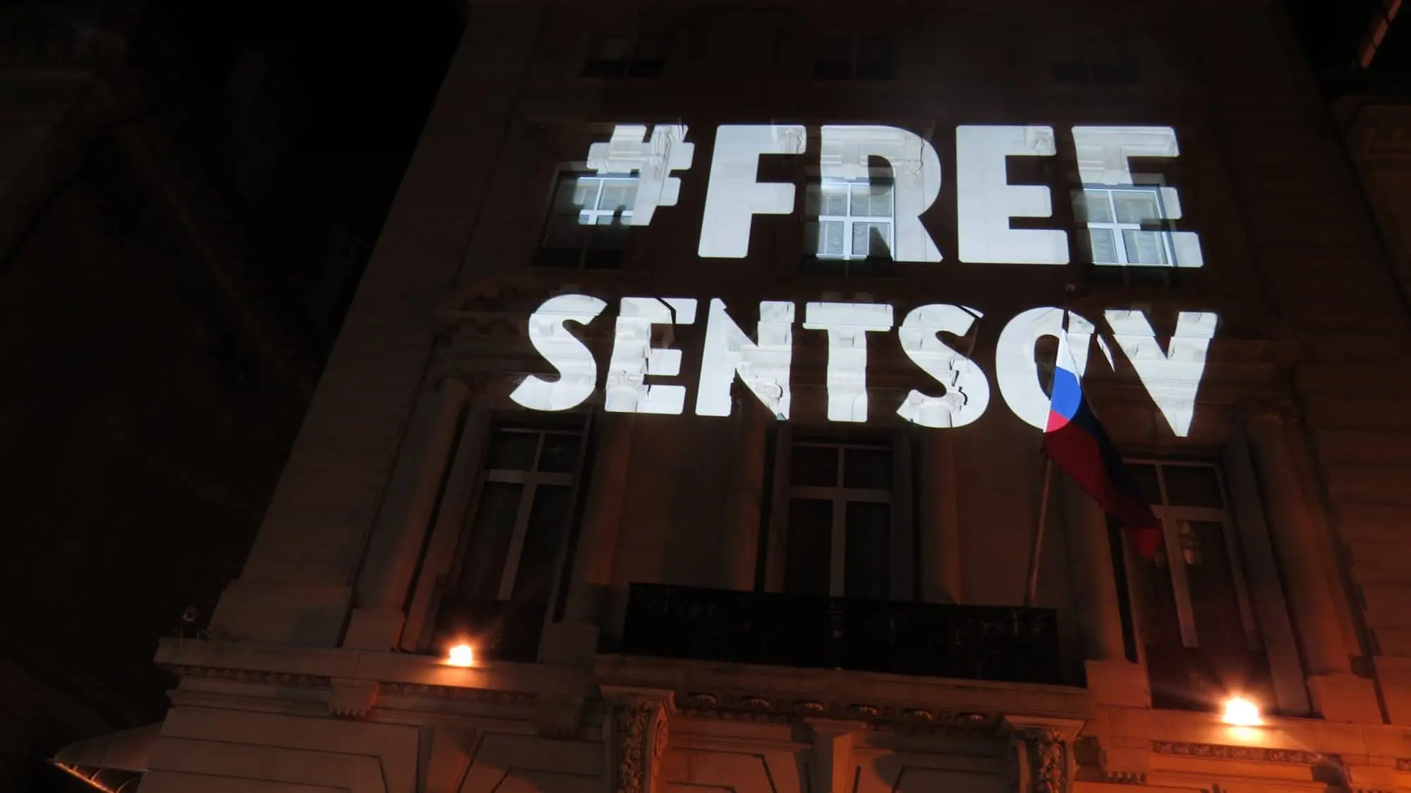Large white text #FREE SENTSOV is projected onto the facade of a building at night. A Russian flag is visible on a balcony below the projection. The building is illuminated by exterior lights.
