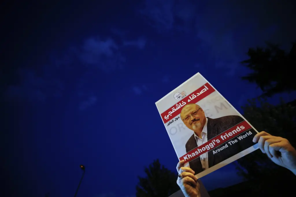A hand holds up a sign with a photo of Jamal Khashoggi and text in English and Arabic, reading Khashoggis friends against a dark blue evening sky.