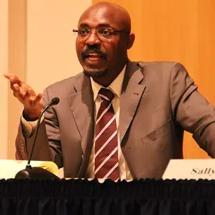 A man in a brown suit and striped tie speaks passionately at a panel, gesturing with his hand. He sits behind a microphone and a black tablecloth, with a name placard partially visible.