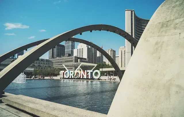 Nathan Phillips Square in Toronto with the iconic TORONTO sign, large arches over a reflecting pool, modern skyscrapers, and a bright blue sky in the background.