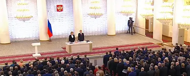 A formal ceremony with officials and guests in a grand hall decorated with chandeliers, a Russian flag, and the state emblem. Two people stand at a podium facing the audience.