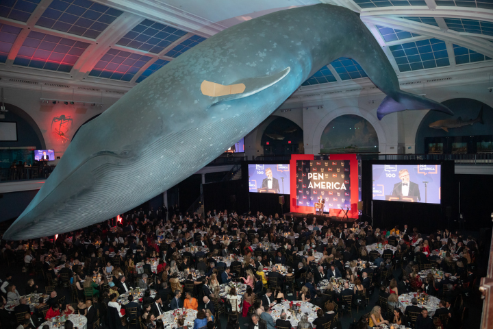 A large indoor event with many seated guests under a life-sized blue whale model, featuring a stage with PEN America signage and large screens displaying a speaker.