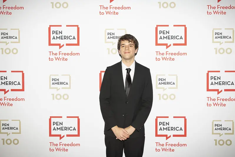 A young man in a black suit and tie stands in front of a step-and-repeat banner with the PEN America logo and the slogan “The Freedom to Write” at a formal event.