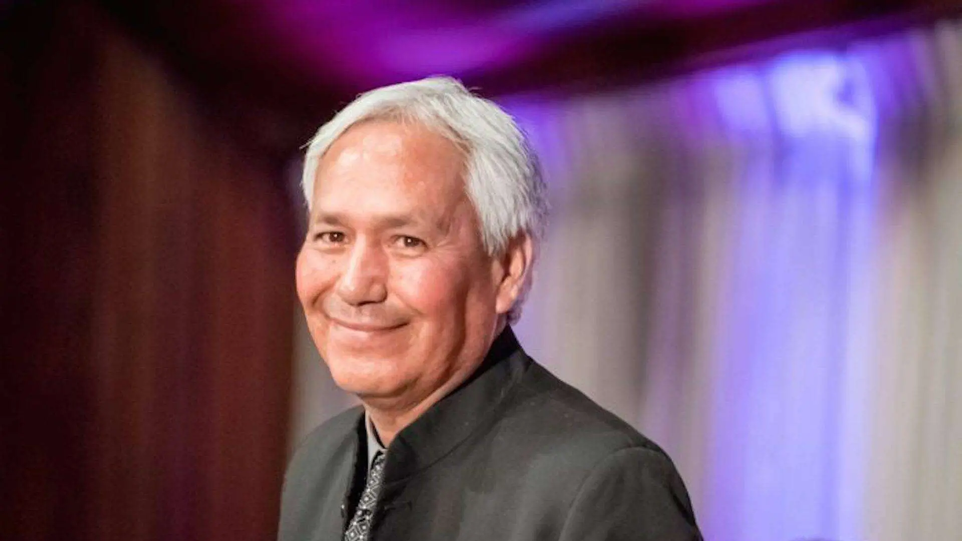 An older man with short white hair smiles while wearing a dark suit and patterned tie, standing in front of softly lit, purple and white draped curtains.