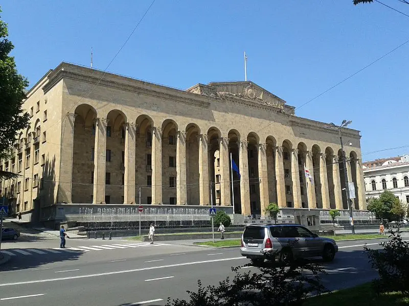 Large, rectangular neoclassical building with tall columns and arched windows, two flags at the entrance, and cars parked on the street in front. Bright blue sky and some trees surround the area.