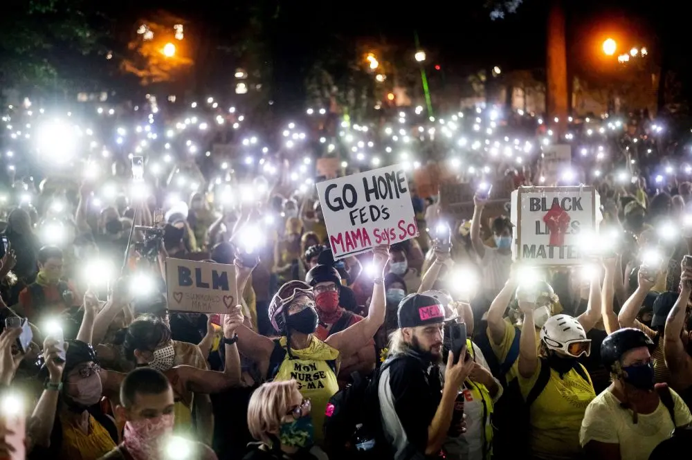 A large crowd at a nighttime protest holds up phones with flashlights on. Visible signs read “GO HOME FEDS MAMA SAYS SO,” “BLM,” and “BLACK LIVES MATTER.” Many people wear masks and yellow clothing.