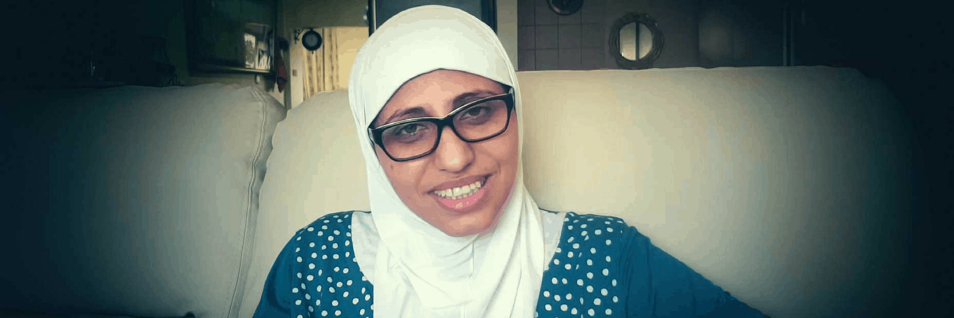 A woman wearing glasses and a white hijab smiles while sitting on a light-colored sofa, with a room and wall decorations visible in the background.