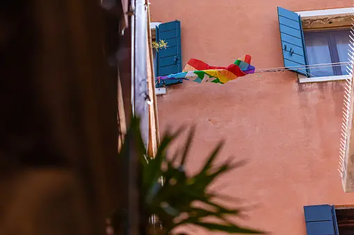 A rainbow flag hangs on a clothesline between two open blue-shuttered windows on a peach-colored building, with green leaves blurred in the foreground.