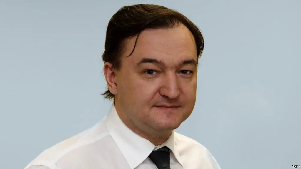 A man with short dark hair wearing a white shirt and dark tie is looking at the camera with a slight smile, posed against a plain light blue background.