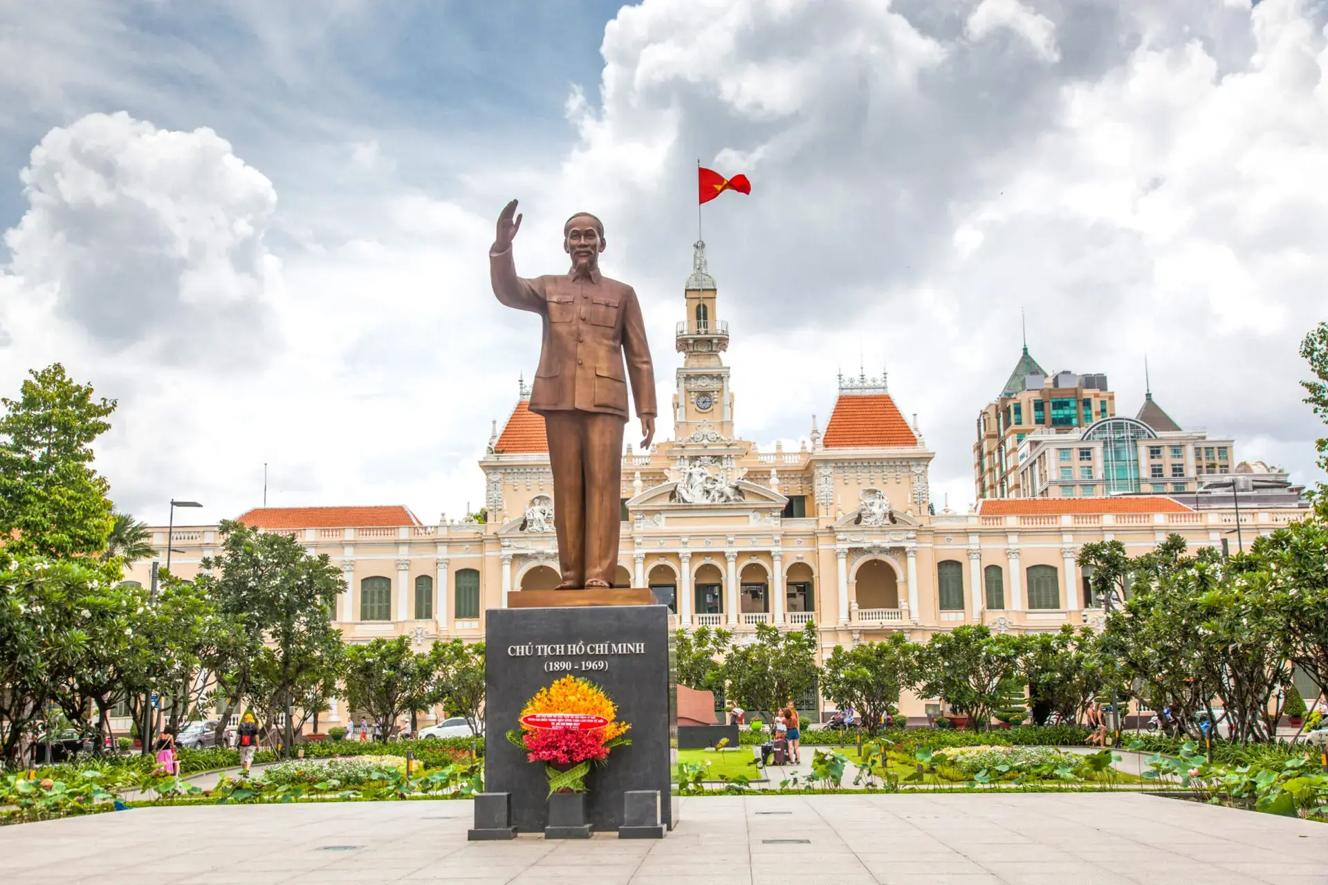 A bronze statue of Ho Chi Minh stands in front of the Ho Chi Minh City Hall, with a Vietnamese flag flying atop the building and people walking in the gardens below a cloudy sky.