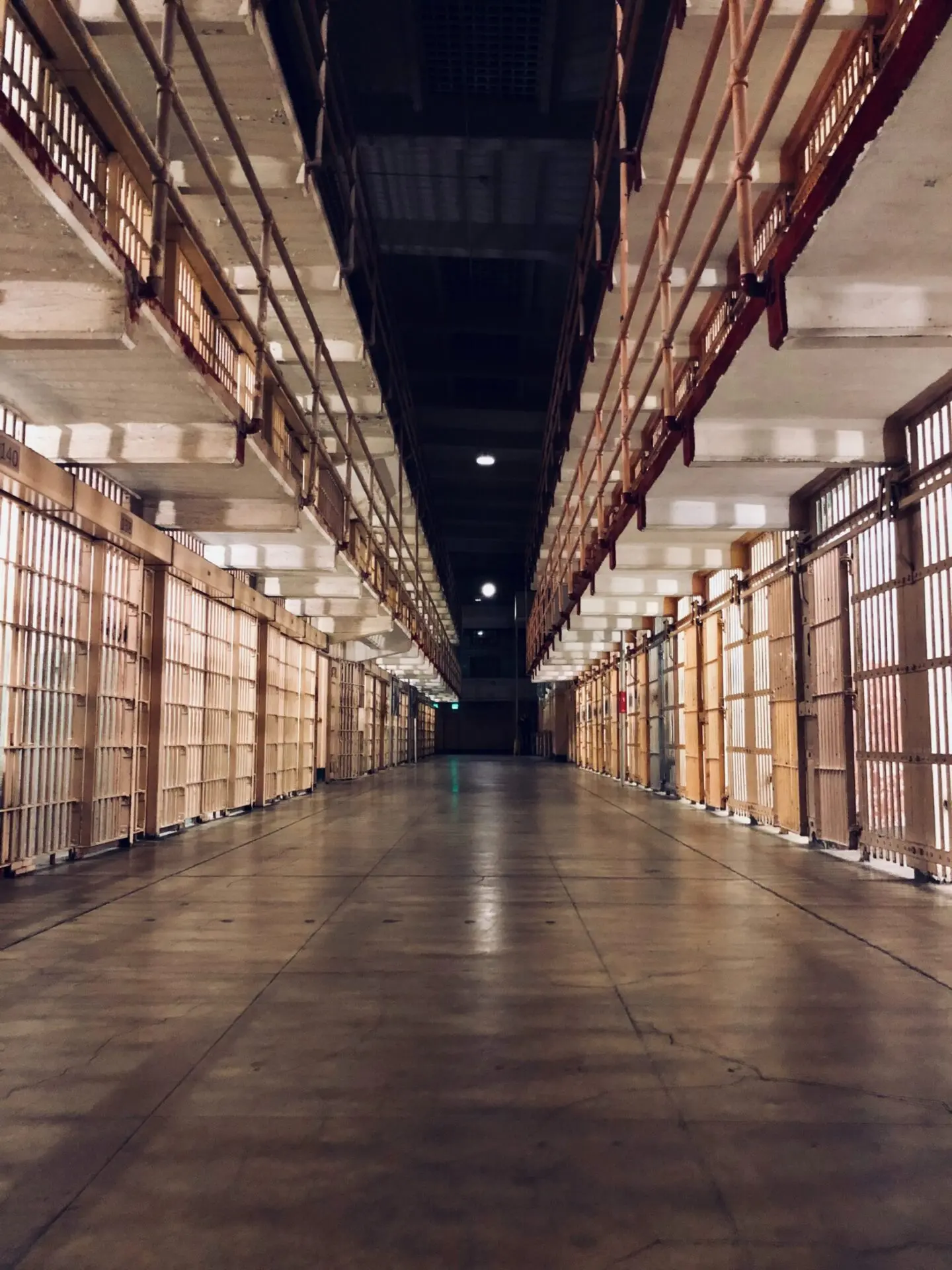 A long, empty hallway in a prison with metal cell doors on both sides and two levels of walkways above, all lit by overhead lights.