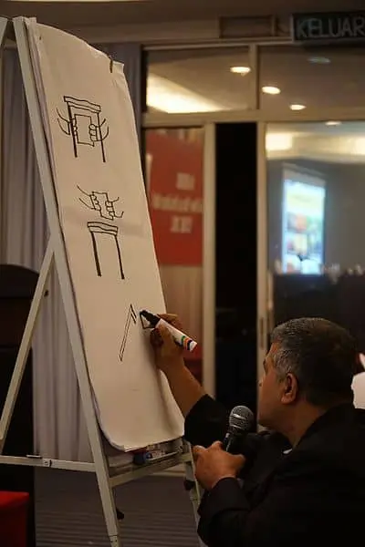 A person holding a microphone and drawing simple, abstract structures with a marker on a large whiteboard at an indoor event. The drawings resemble frames or gates.