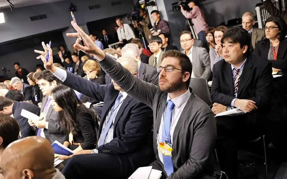 A group of journalists sit in a conference room; two men in the front row raise their hands, holding pens, eager to ask questions. Others listen attentively or take notes.