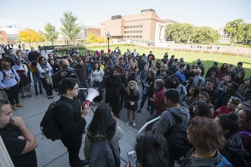 A large group of students gathers outdoors on a college campus, listening to a person speaking through a megaphone about PEN America. The crowd stands on a sidewalk, with campus buildings and green lawns visible in the background.