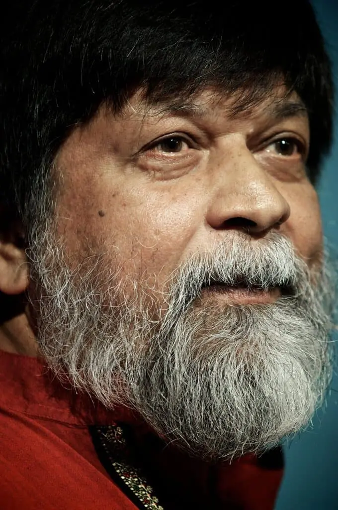 A close-up of an older man with a thick gray and white beard and mustache, wearing a red garment with intricate detail, looking slightly upward with a gentle expression.