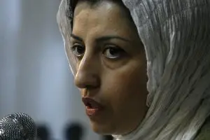 A woman wearing a light-colored headscarf speaks into a microphone, shown in close-up from the side. Her expression appears serious and focused. The background is softly blurred.