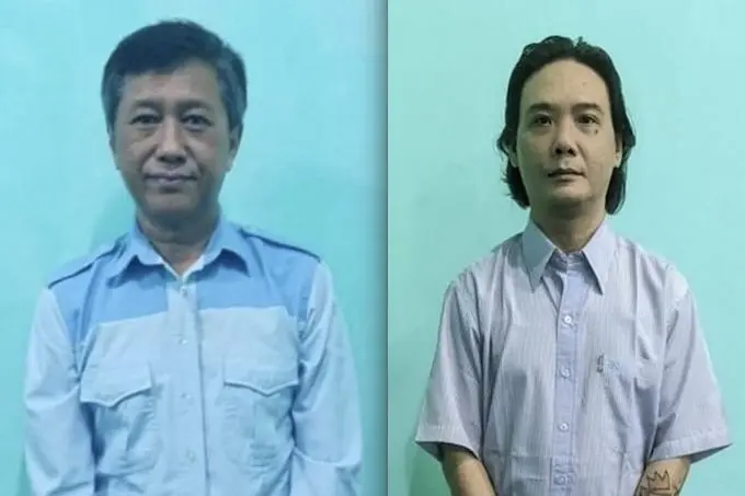 Two men stand side by side against a plain teal background, both wearing collared shirts. The man on the left has short dark hair and a light blue shirt; the man on the right has longer hair and a light purple shirt.