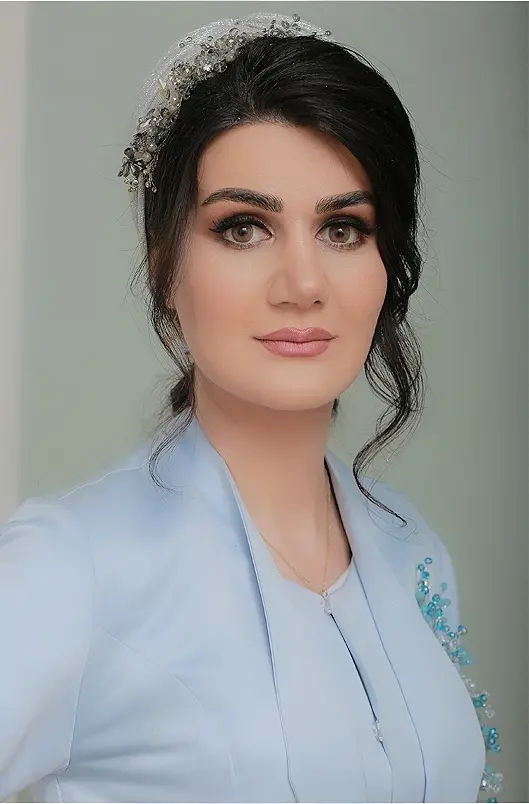 A woman with dark hair styled in loose waves, wearing a light blue outfit with decorative beads, and a silver hair accessory, poses in front of a neutral background. She has striking makeup and a calm expression.