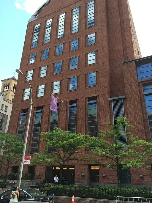 A tall red-brick building with large windows and a purple flag hanging above the entrance. Several trees line the sidewalk, and people and cars are visible on the street in front.