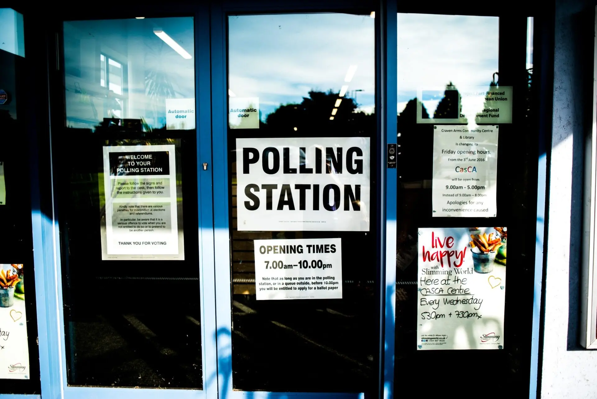 Glass doors display several signs, including a large one reading “POLLING STATION” and another listing opening times as 7:00 a.m. to 10:00 p.m.; other posters provide local information and announcements.