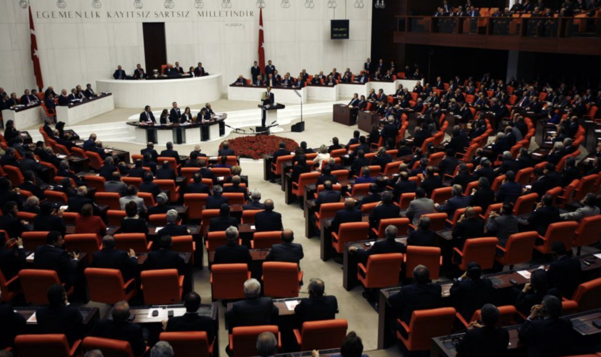 A large assembly hall filled with people seated in orange chairs, listening to a speaker at a central podium. The space is formal, with Turkish flags and a circular arrangement facing the speaker.