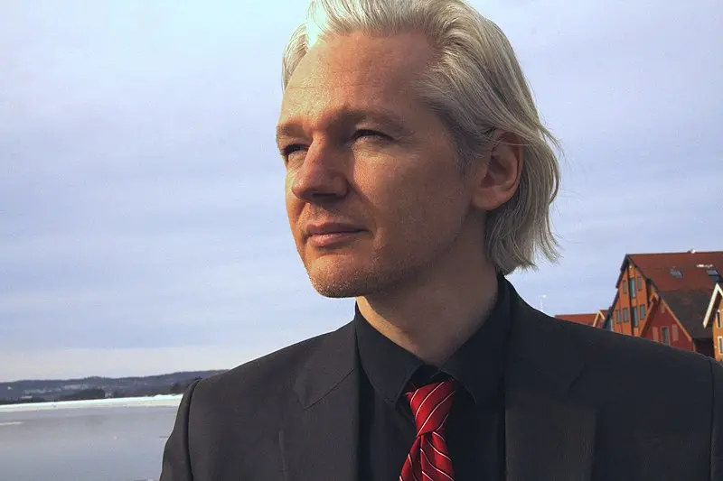 A man with pale skin and shoulder-length blond hair stands outdoors, wearing a black suit, black shirt, and red striped tie. He looks thoughtfully into the distance; houses and a body of water appear in the background.
