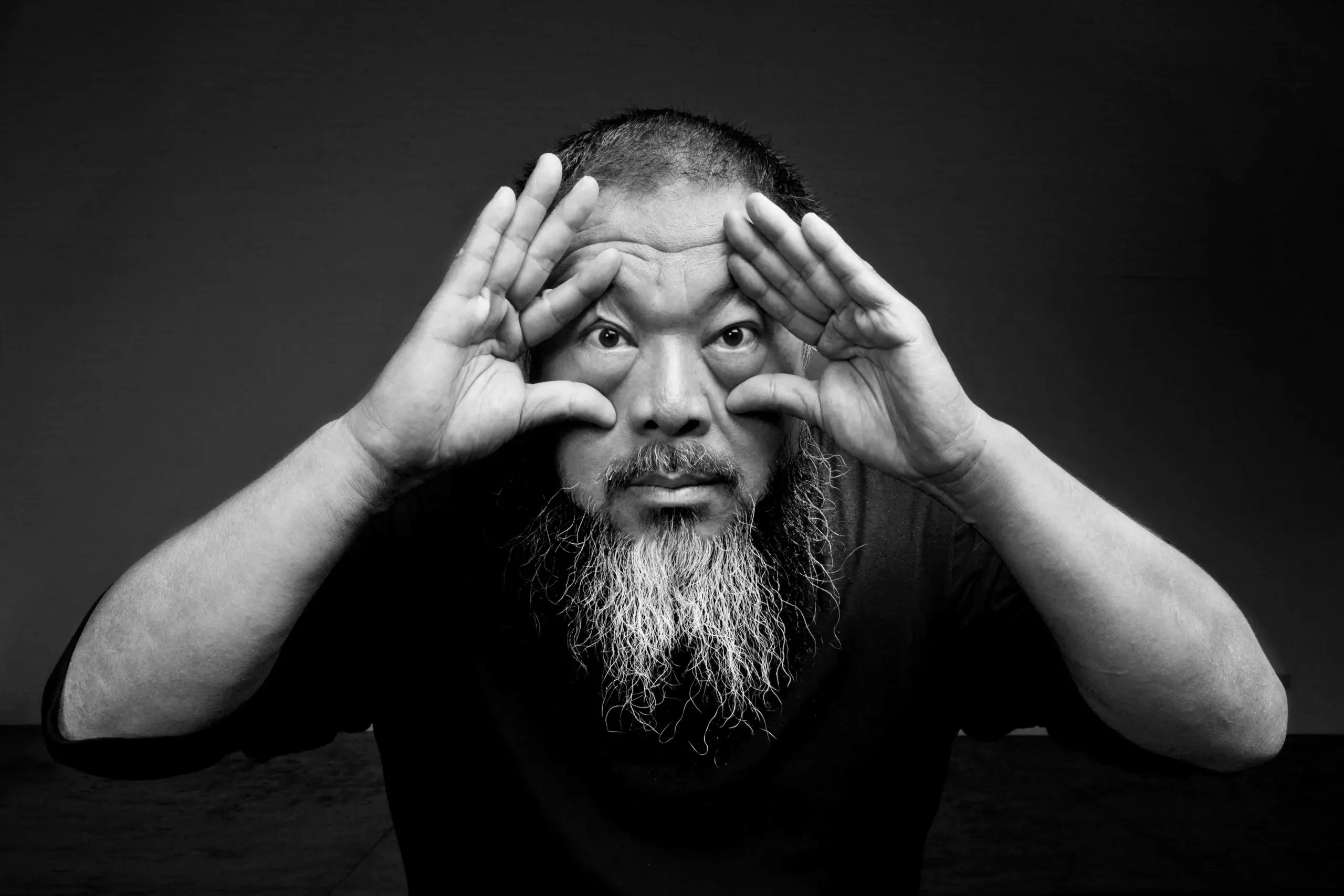 A man with a long beard holds his hands around his eyes, mimicking binoculars, and looks directly at the camera. The photo is black and white with a dark background.