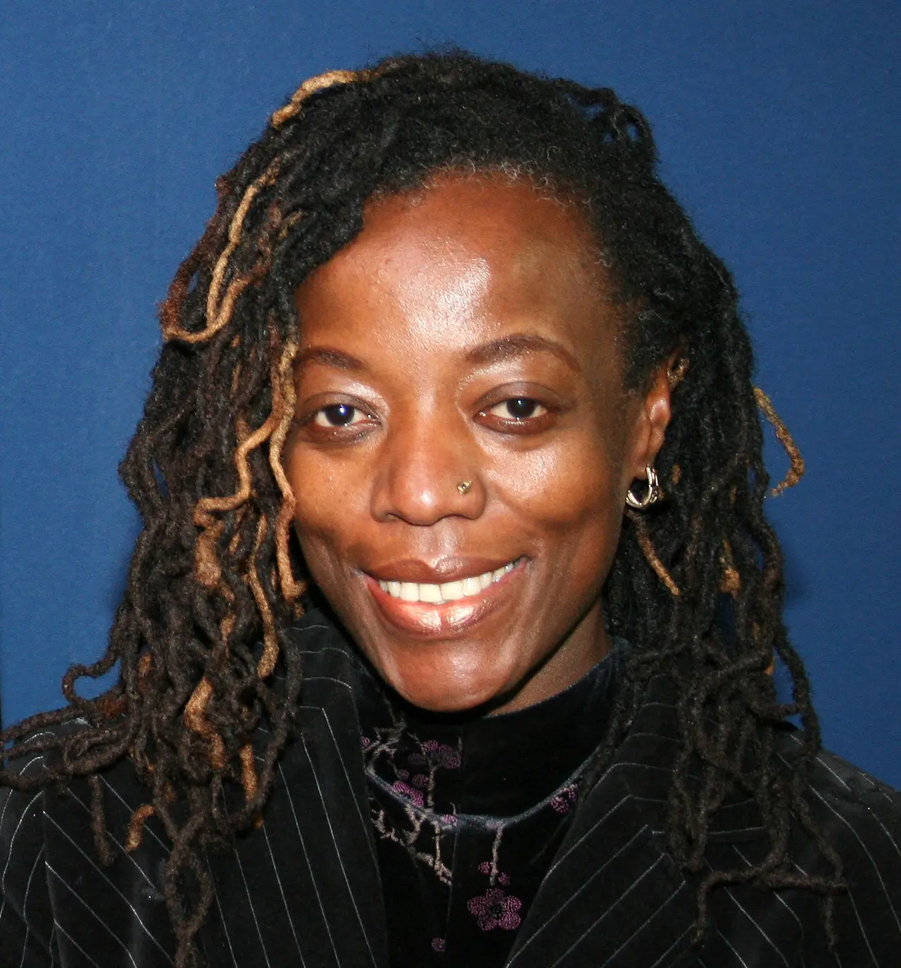 A person with long, dark, and light brown dreadlocks smiles at the camera. They are wearing a dark, pinstriped jacket and a nose ring, with a blue background behind them.