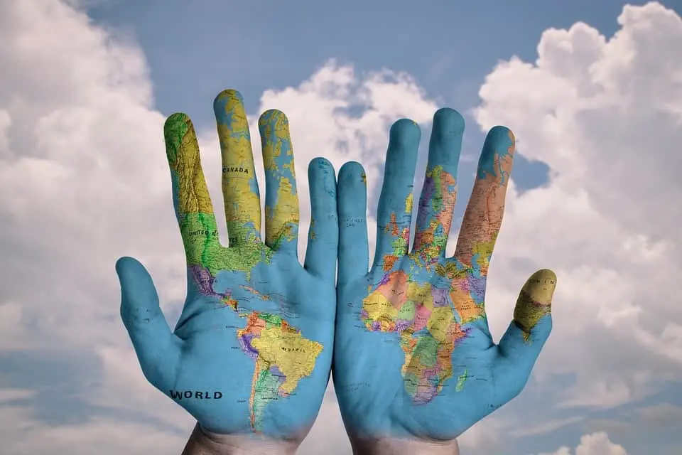 Two open hands are held up against a cloudy sky, with a world map painted on the palms and fingers, displaying continents and countries across both hands.