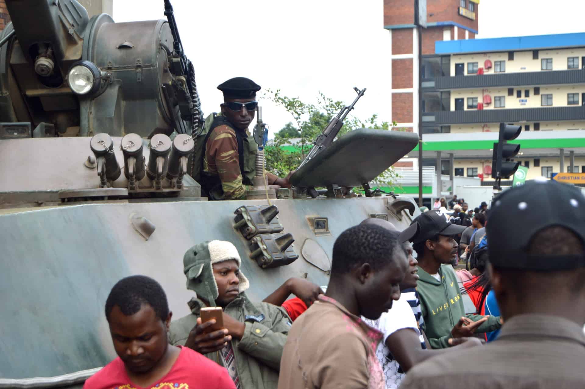 A soldier in uniform sits atop an armored vehicle amid a crowd of people on a city street, with buildings visible in the background. Some people are looking at the vehicle, while others are using their phones.