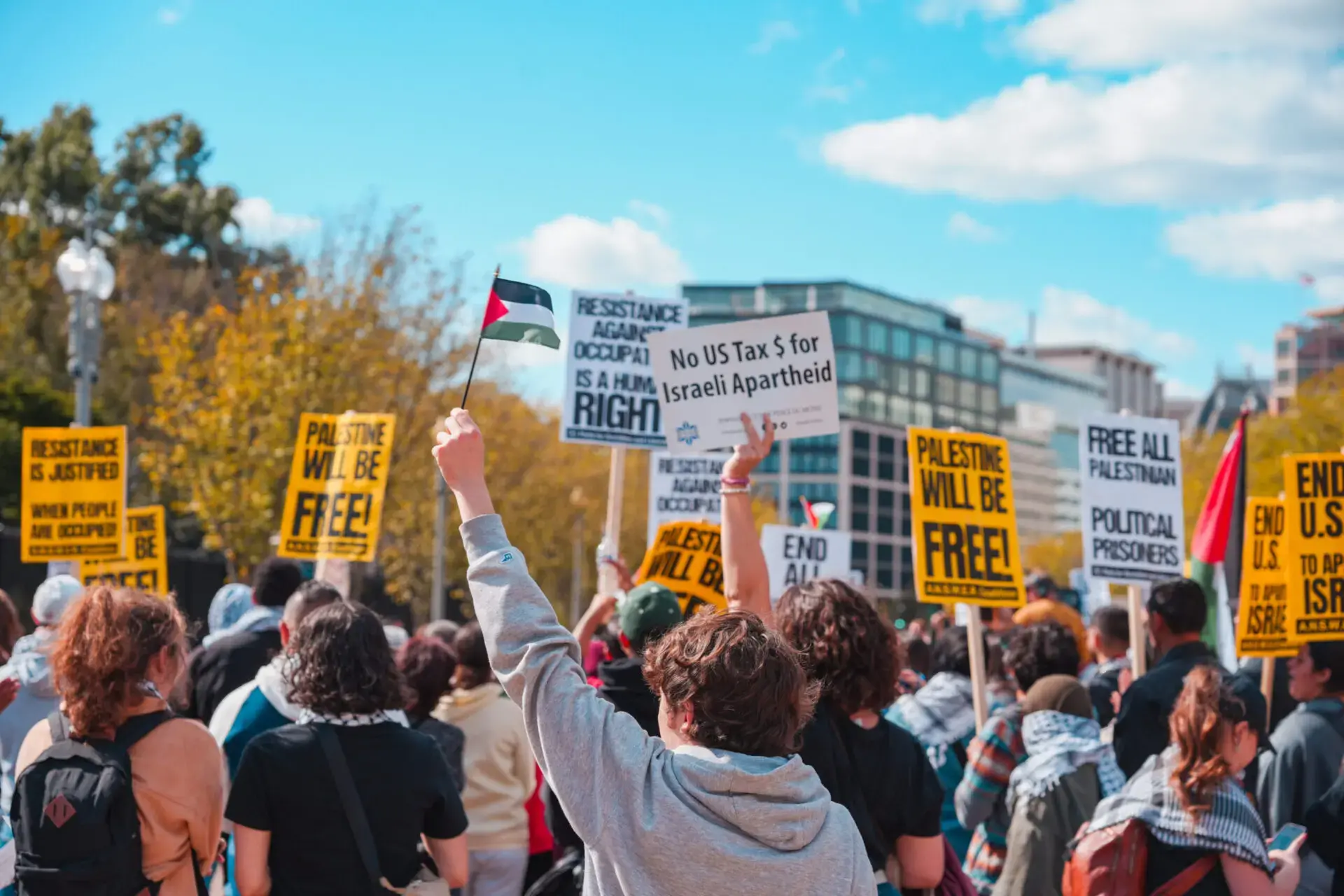 A crowd of protesters hold signs with messages about ending Israeli occupation and freeing Palestine. One person waves a Palestinian flag, and the scene takes place outdoors with buildings and trees in the background.