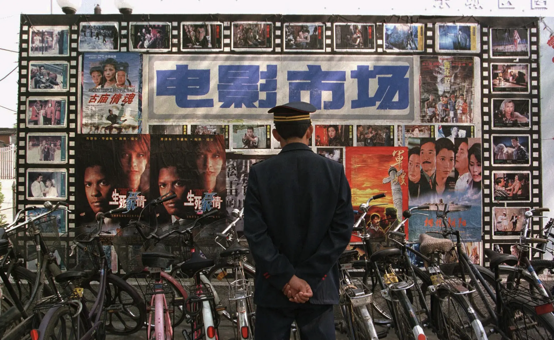 A person in uniform stands with hands behind their back, facing a wall covered with Chinese movie posters and a large sign, surrounded by parked bicycles.