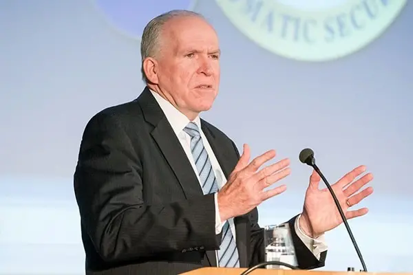 An older man in a suit and striped tie speaks at a podium with a microphone, gesturing with his hands. A blurred official seal is visible in the background.