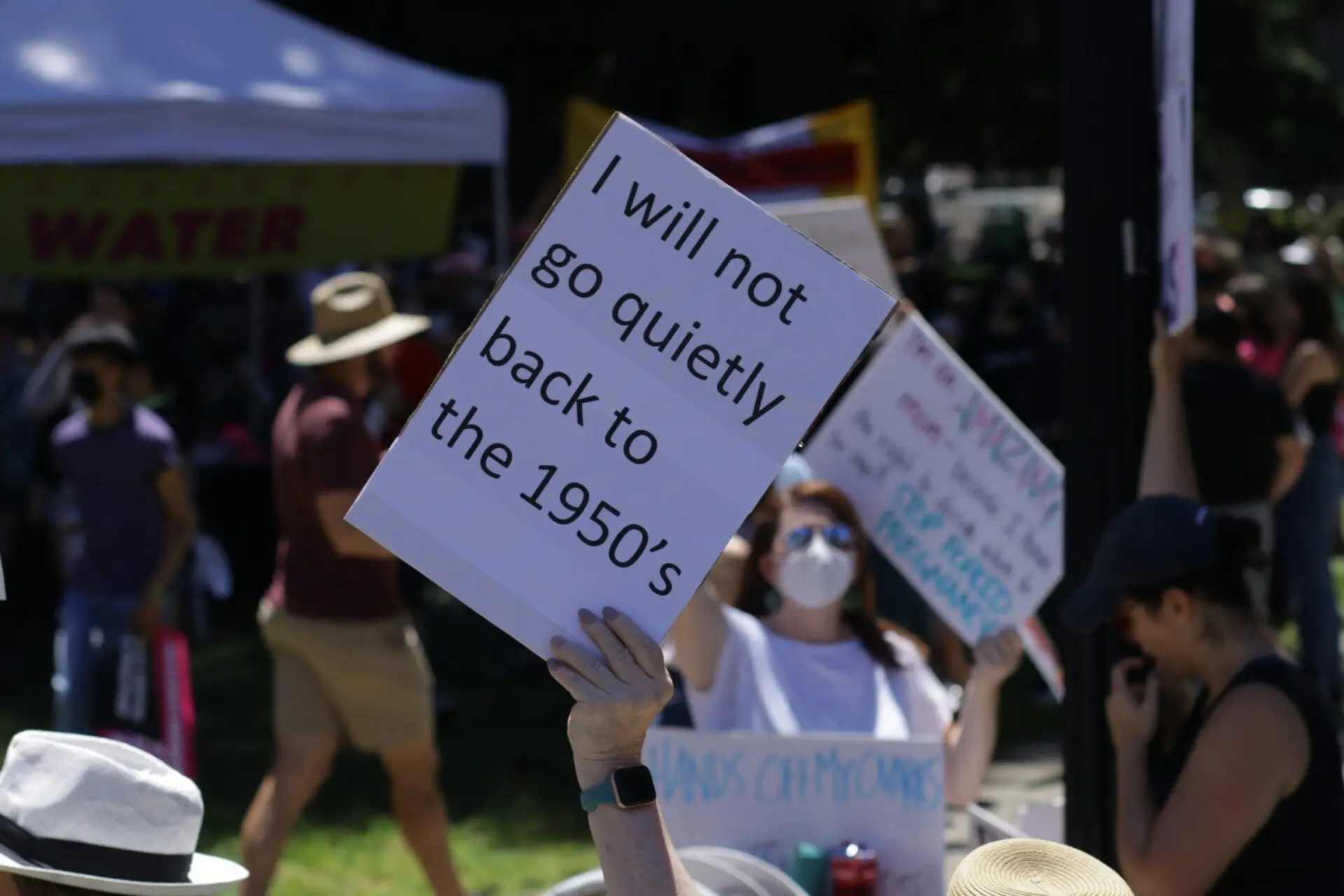 A person at a protest holds a large sign reading, I will not go quietly back to the 1950s. Other people with signs and wearing hats or masks are visible in the background.