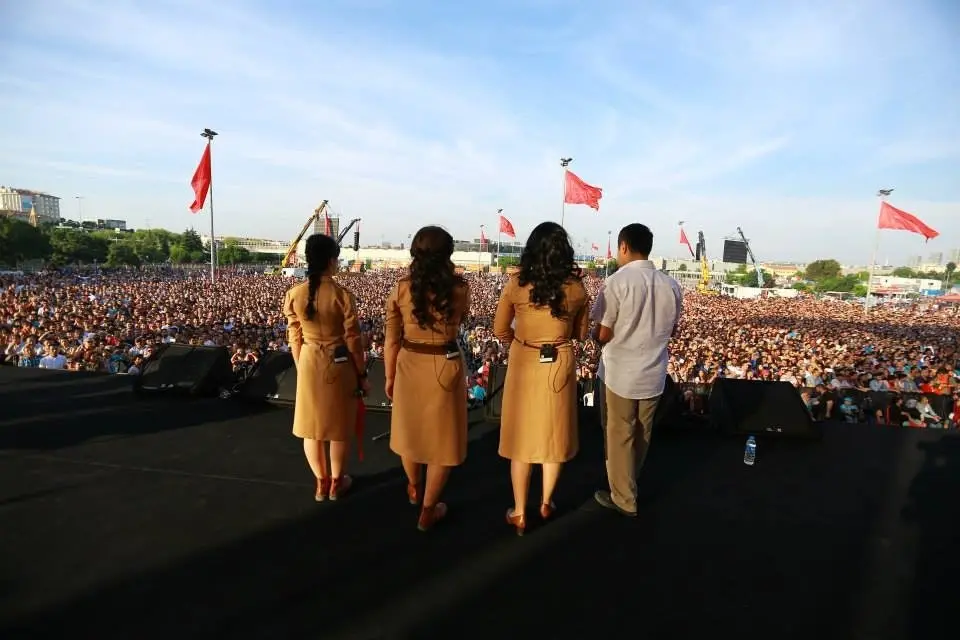Four people, three women in matching brown dresses and one man in a light shirt, stand on an outdoor stage facing a large crowd under a blue sky with red flags visible in the background.