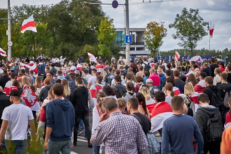A large crowd of people with red and white flags walk down a street during a protest or demonstration, surrounded by trees and street signs in an urban setting.