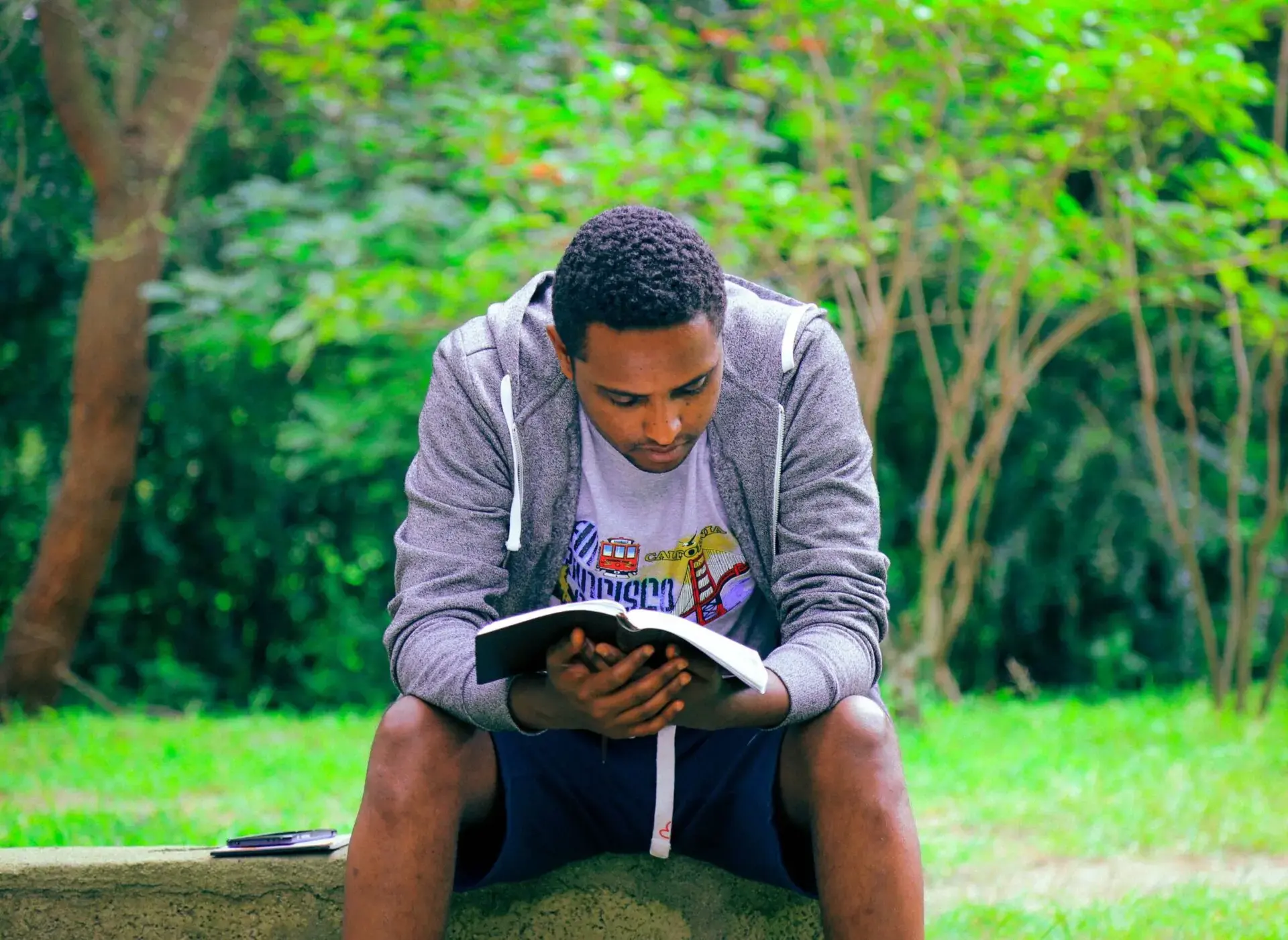 A young man sits on a stone bench outdoors, absorbed in reading a book, with green trees and grass in the background. He wears a gray hoodie, shorts, and a white T-shirt.