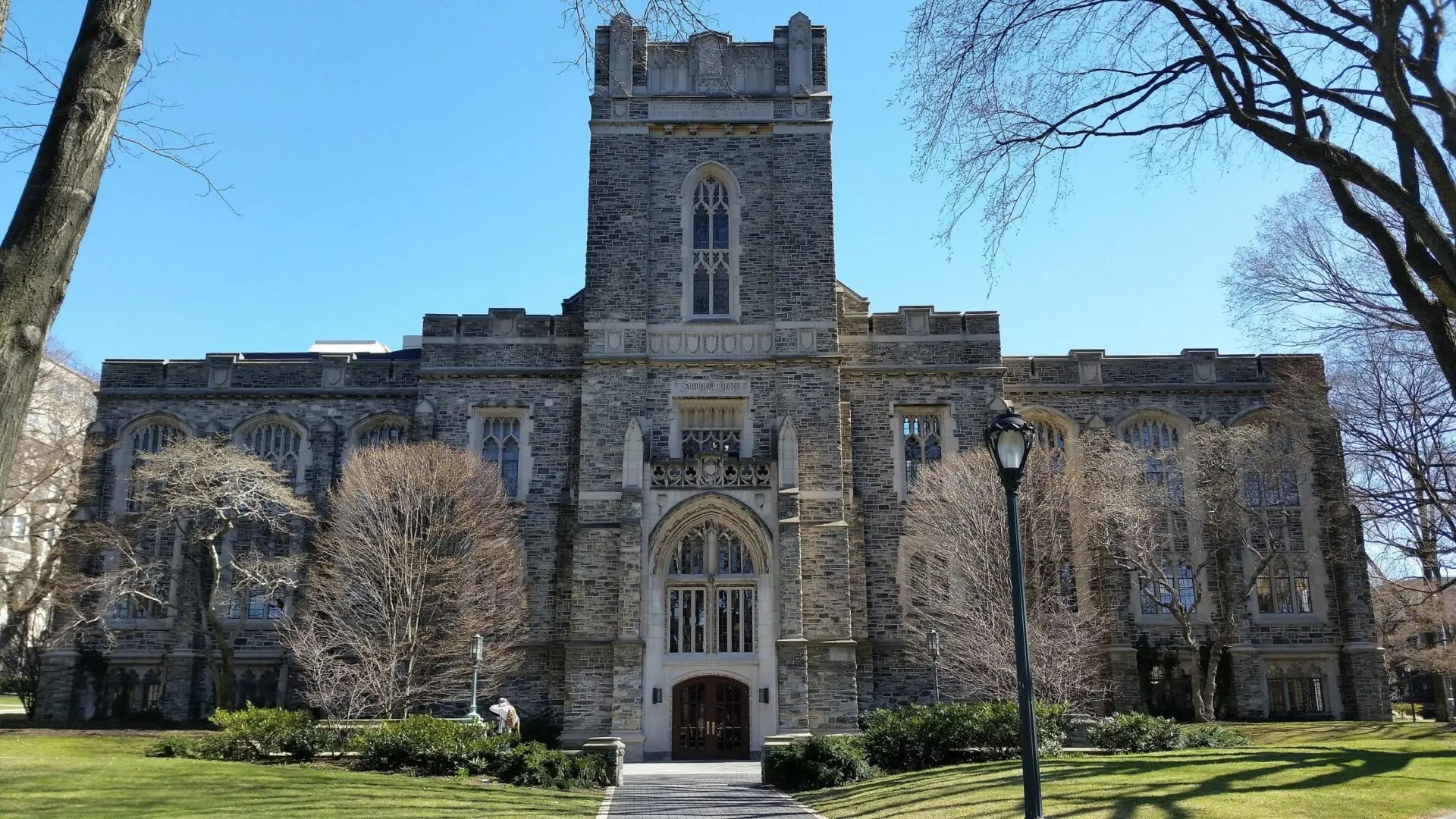 A large stone building with tall arched windows and a central tower, surrounded by trees and a lawn, under a clear blue sky.