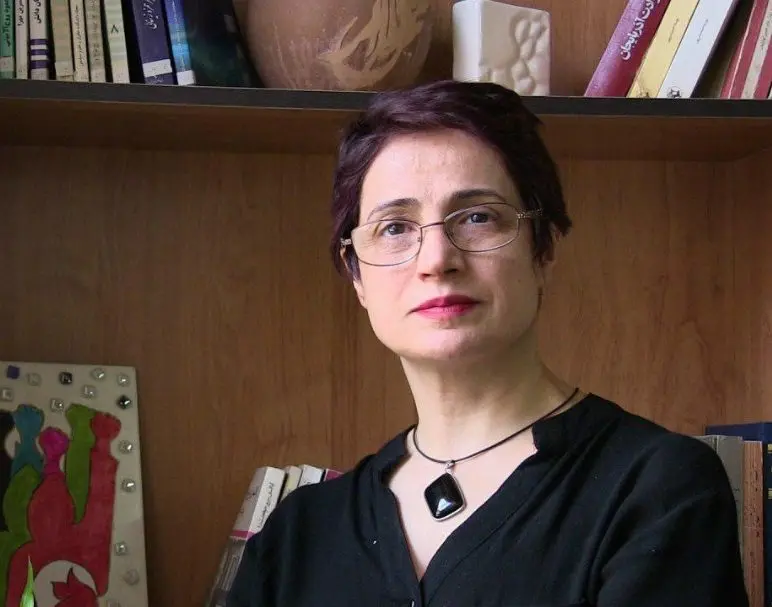 A woman with short dark hair and glasses, wearing a black top and a pendant necklace, stands in front of a wooden bookshelf filled with books and decorative items, looking directly at the camera.