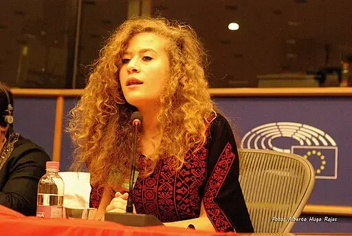 A woman with long, curly hair speaks into a microphone at a panel, seated in front of seats and a blue backdrop featuring an EU emblem. There is a water bottle and papers on the table in front of her.