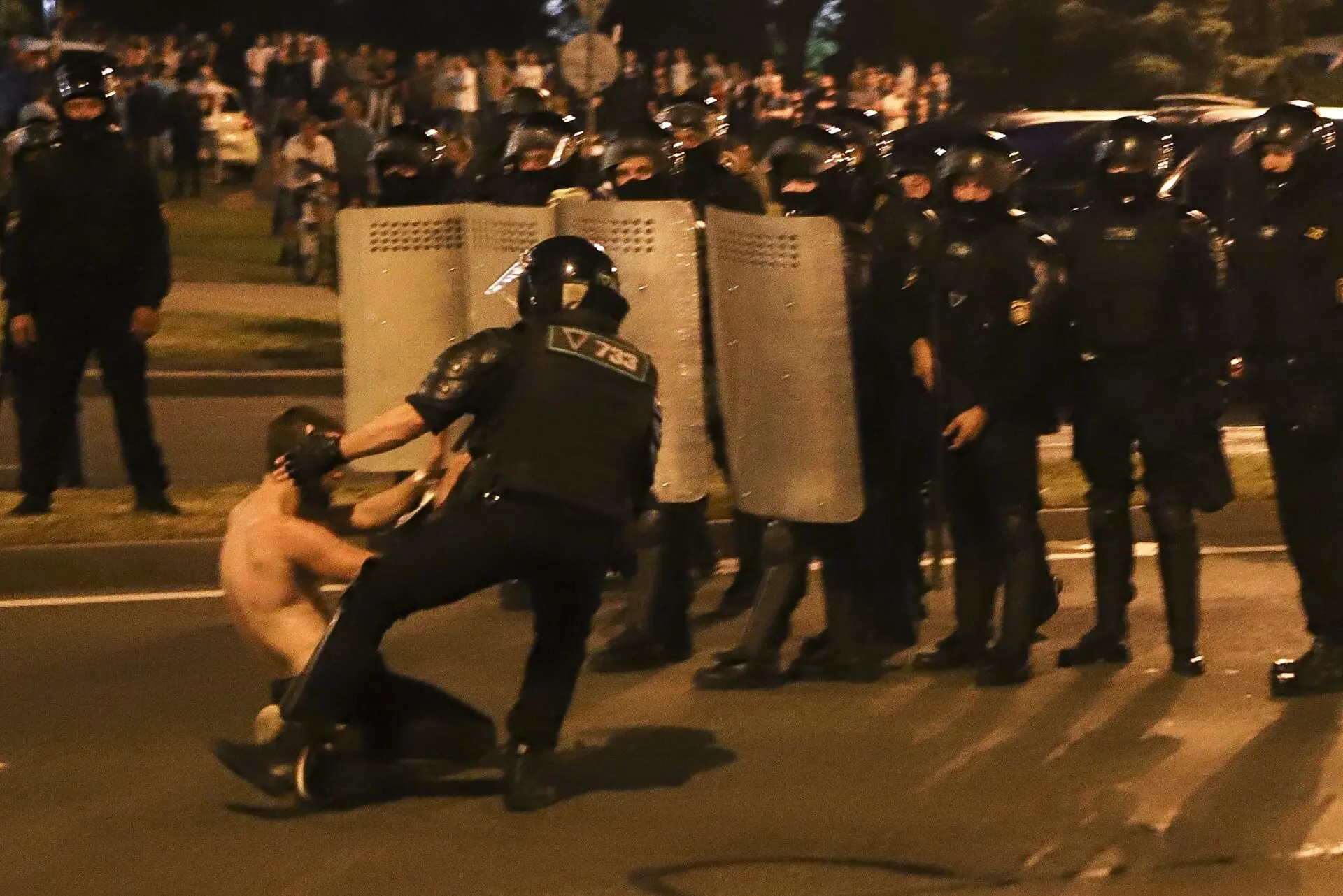 A police officer pulls a shirtless person on the ground during a nighttime protest, while a line of officers with riot shields stands behind them. A crowd watches from the background.
