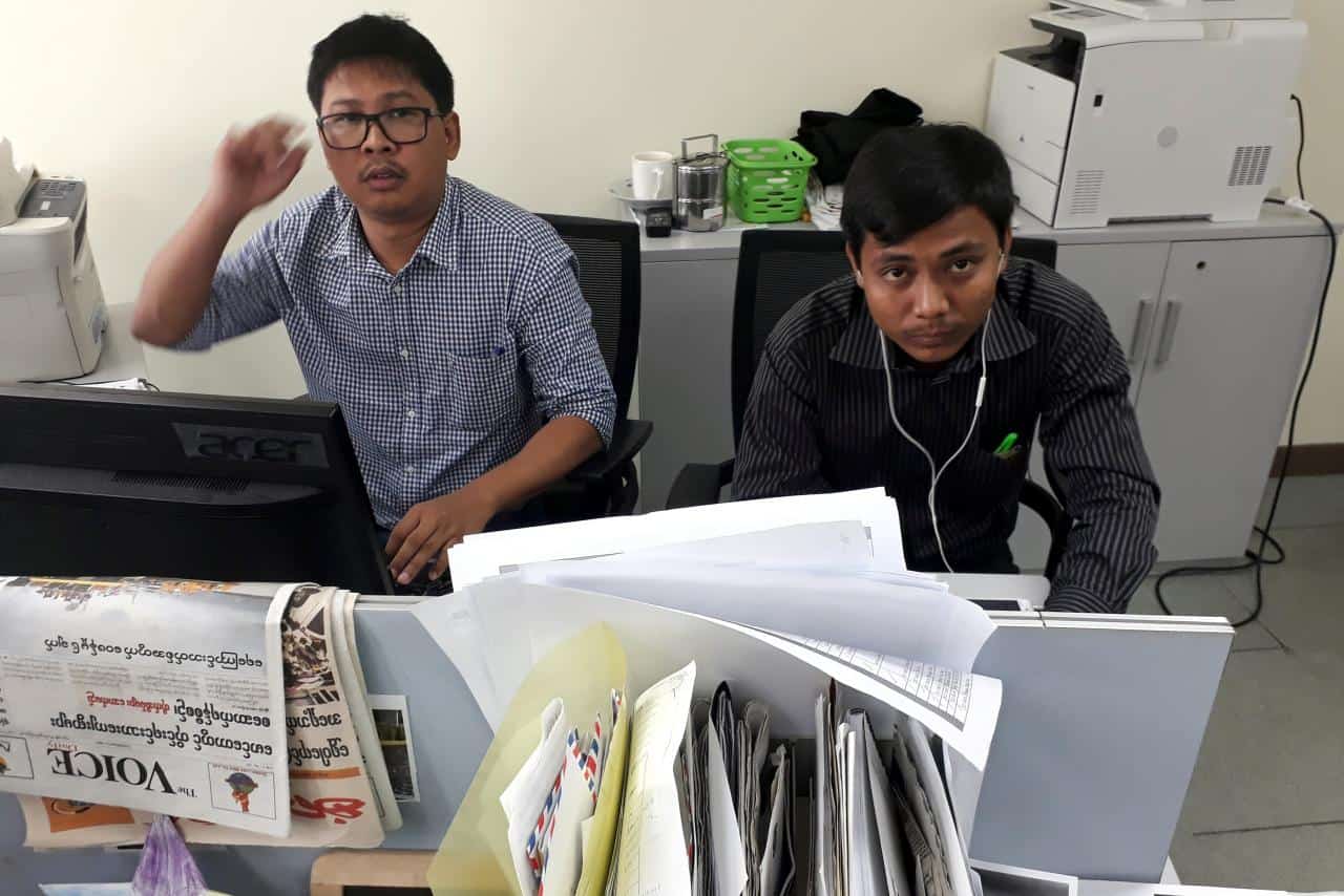 Two men sit at desks in an office, looking up toward the camera. Papers and folders cover the front desk, and office equipment like a printer and baskets are visible in the background.