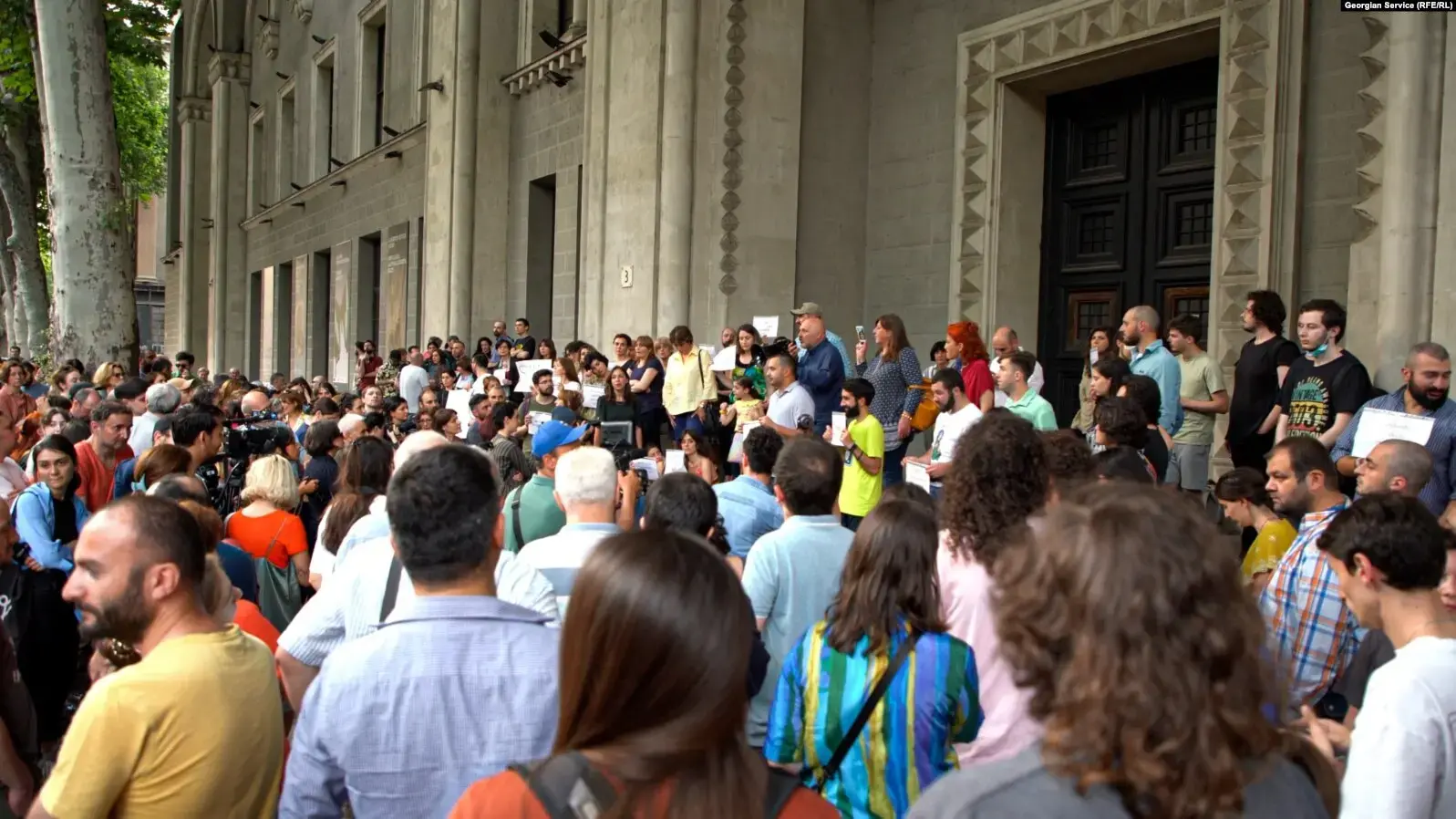 A large crowd gathers outside a historic building with columns and arched windows. People are standing close together, some appear to be listening to speakers or holding signs. Trees line the sidewalk beside the building.